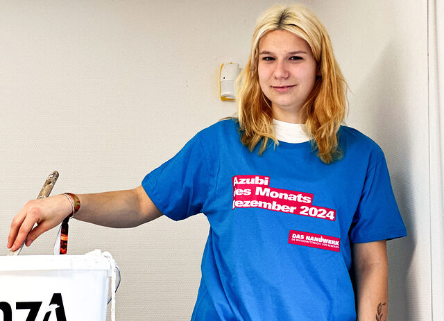 Janine Henke im blauen T-Shirt „Azubi des Monats Dezember 2024“ mit Farbroller und Farbeimer