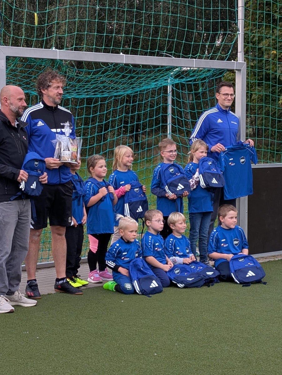 Geförderte Jugend-Fußballmannschaft mit ihren Trainern auf dem Sportplatz, die neue blaue Trikots und Sporttaschen präsentieren